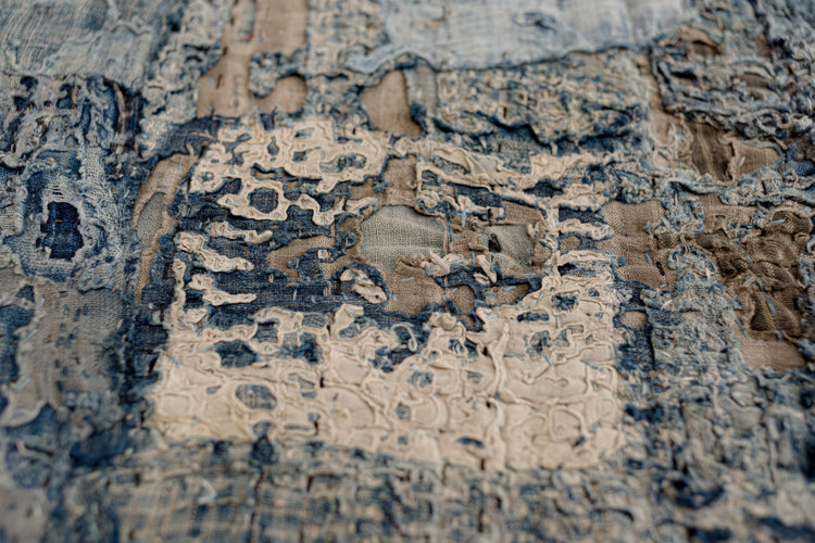 Close-up of a Japanese boro textile showing layered indigo cotton patches hand-stitched together, with frayed edges, visible repairs, and lighter beige fabric exposed beneath worn areas.